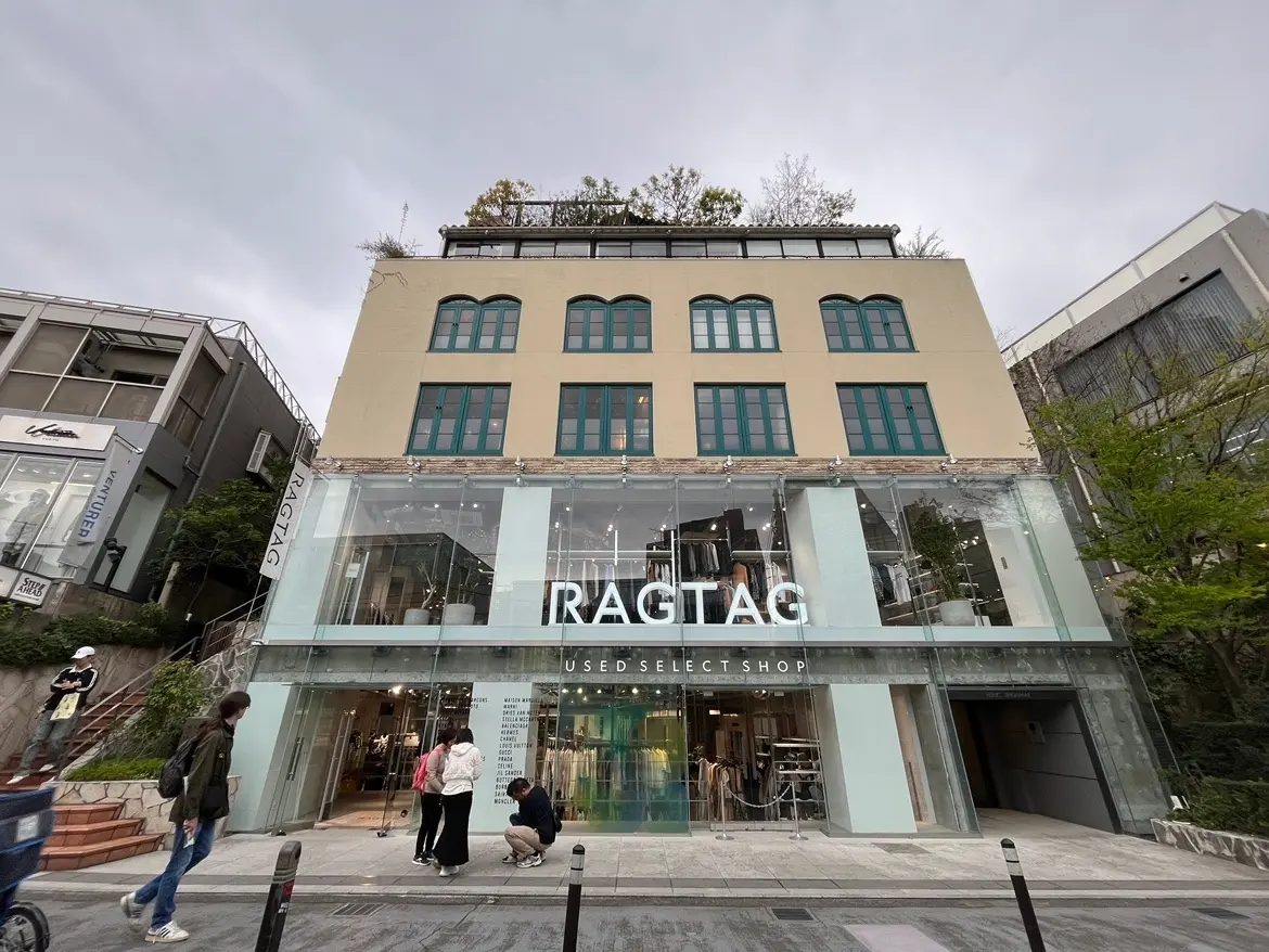- 「RAGTAG」のロゴが印象的なガラス張りの中古セレクトショップの外観、道を歩く人々と周囲の緑が調和した風景