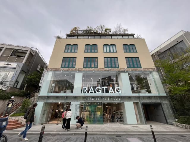 - 「RAGTAG」のロゴが印象的なガラス張りの中古セレクトショップの外観、道を歩く人々と周囲の緑が調和した風景