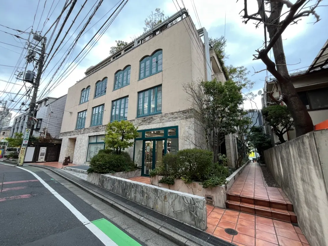 - おしゃれな外観の商業ビル、青い窓枠と植栽、周辺の街並みと交通信号が映る通りの風景