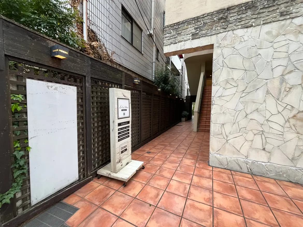 - 建物のエントランスに続くタイル敷きの通路、壁には木製のフェンスと照明、奥に階段が見える外観の写真