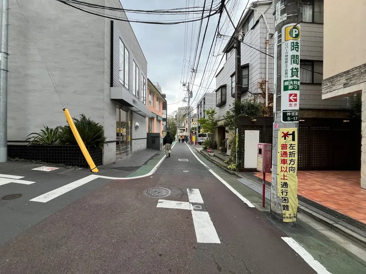 - 地元の商業街の風景、右手に店舗と左折指示の看板があり、舗装された道路に車両通行の案内がある街並み