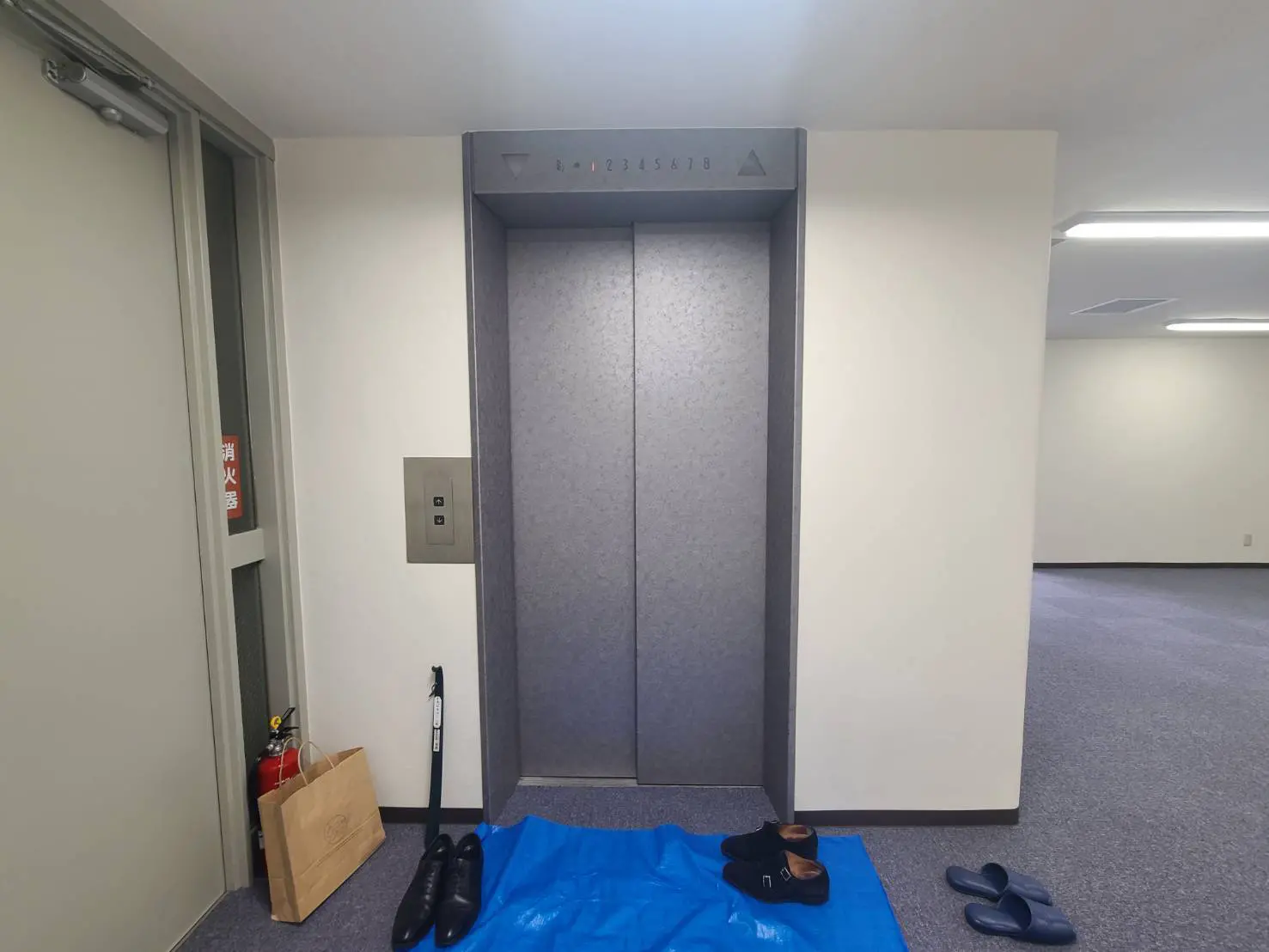 - エレベーターの前に置かれた靴とスリッパ、青いシートの上に立つシンプルな内装のエントランスエリア