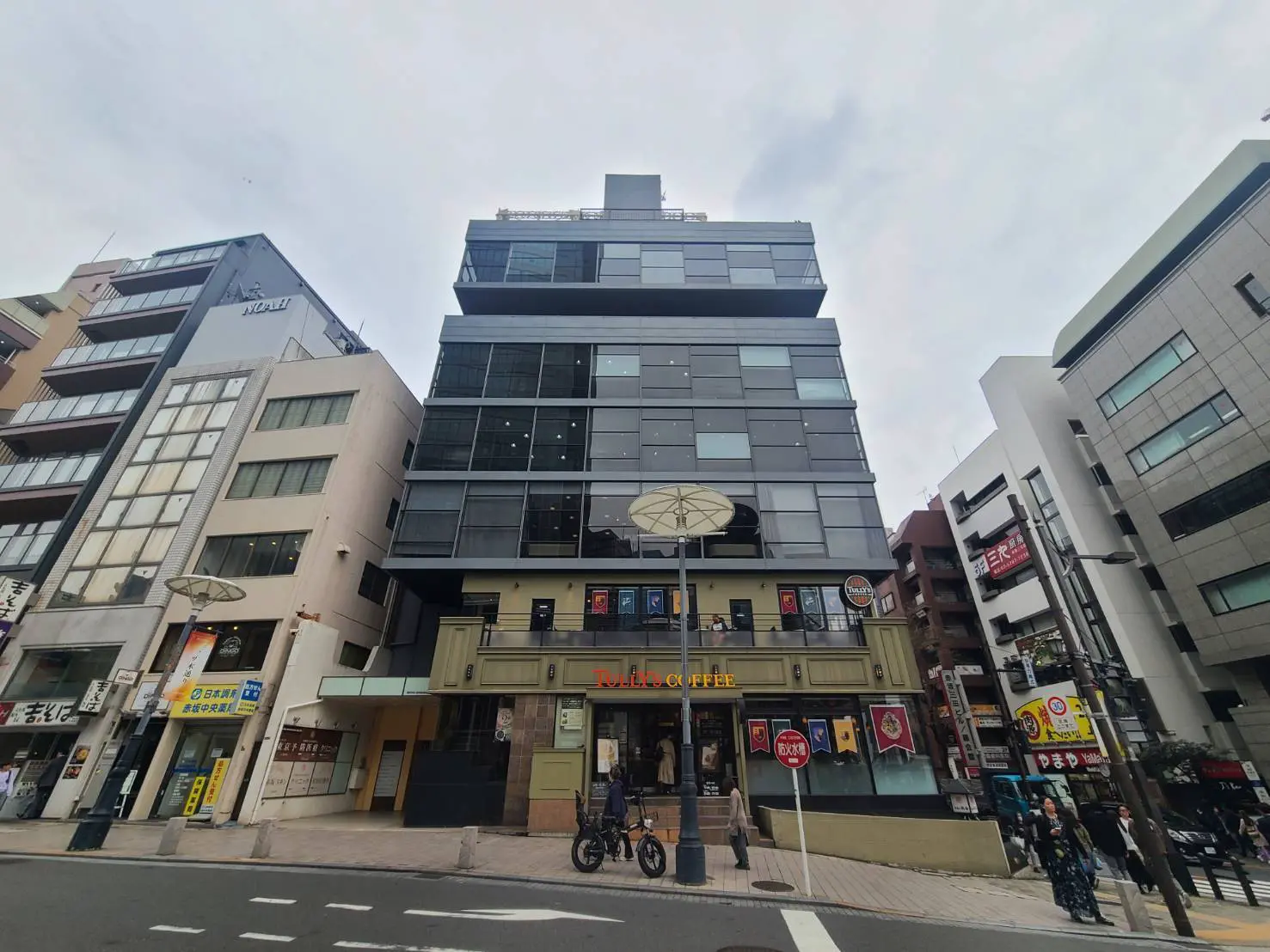 - 現代的なデザインのビルの外観、1階にはカフェがあり、周囲に他の店舗が立ち並んでいる賑やかな通りの風景