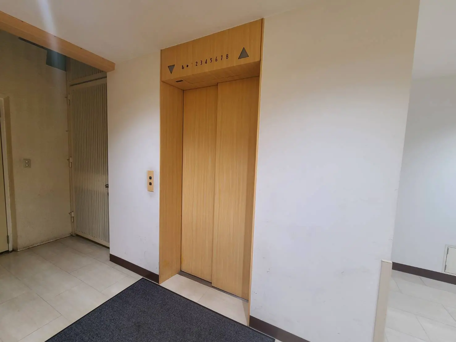 - 木目調のエレベーターの扉がある入口、シンプルで清潔感のある内装、隣にはドアが見えるエントランスエリア