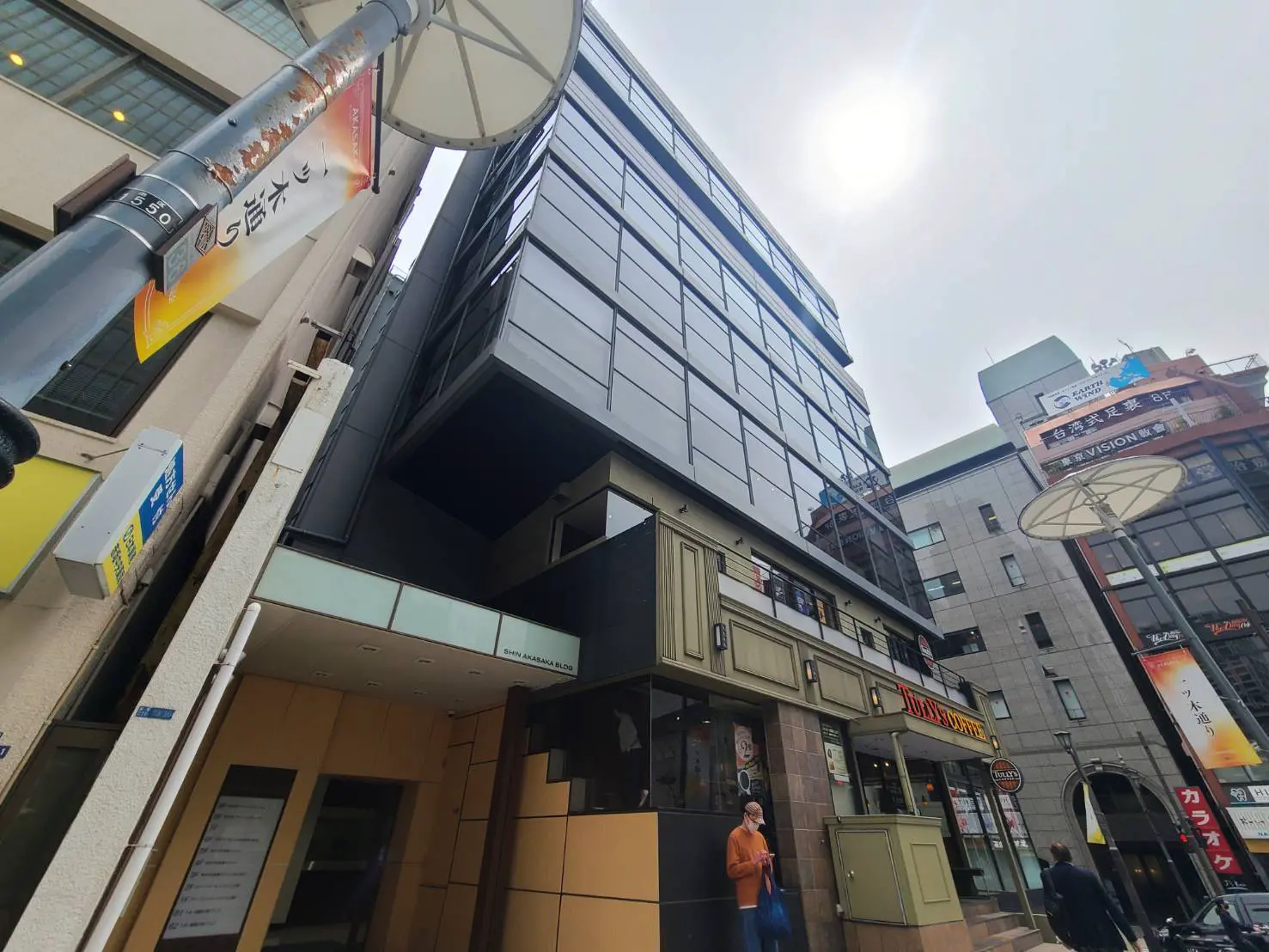 - 現代的な外観のビル、ダークなガラスと明るいエントランス、周囲に店舗が並ぶ賑やかな街並み