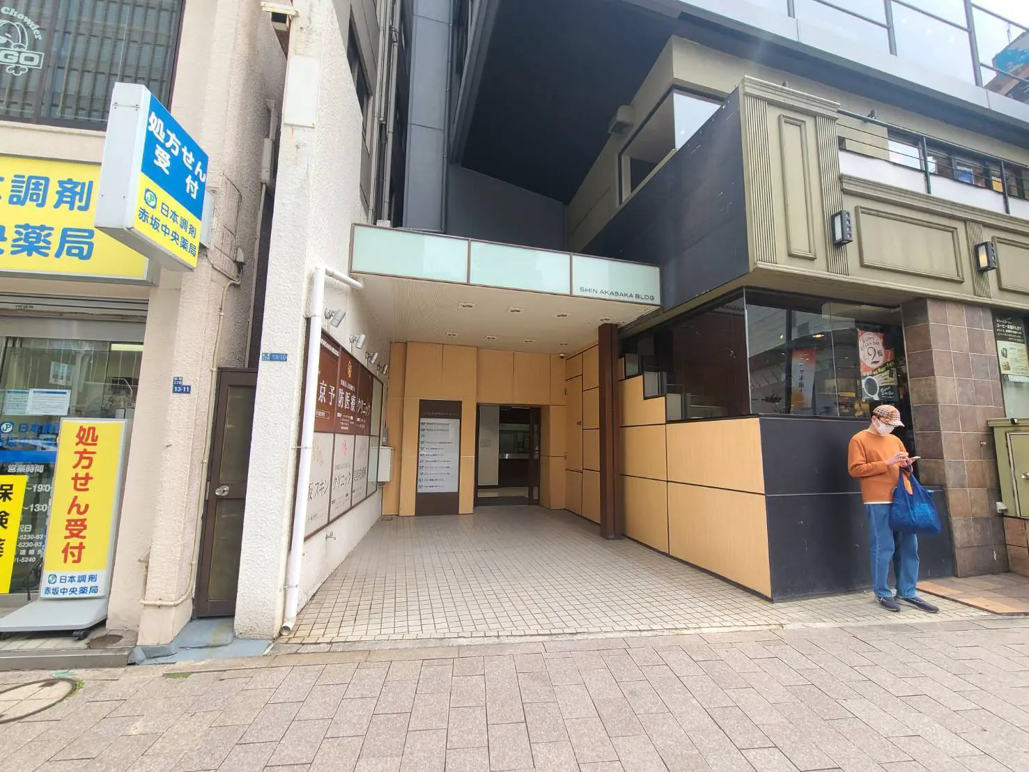 - 商業ビルの入り口、隣に調剤薬局の看板がある街中の風景、スマートフォンを操作する男性が立っている様子