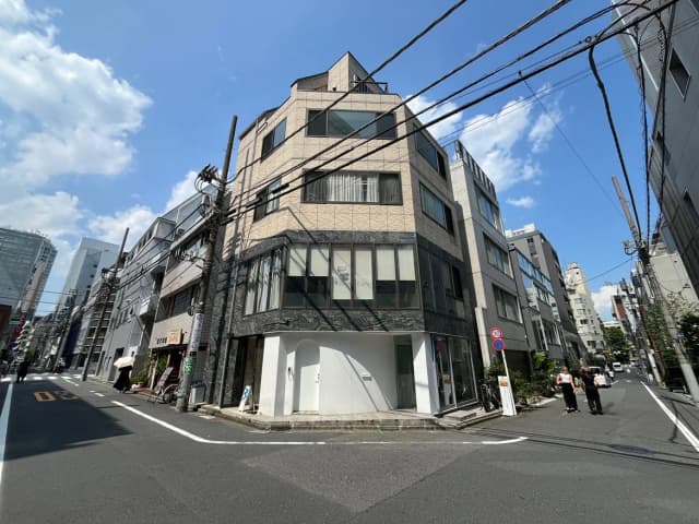 - モダンなデザインのビルと周辺の街並み、青空と白い雲が広がる日中の風景、歩行者が通る賑やかな通りの様子