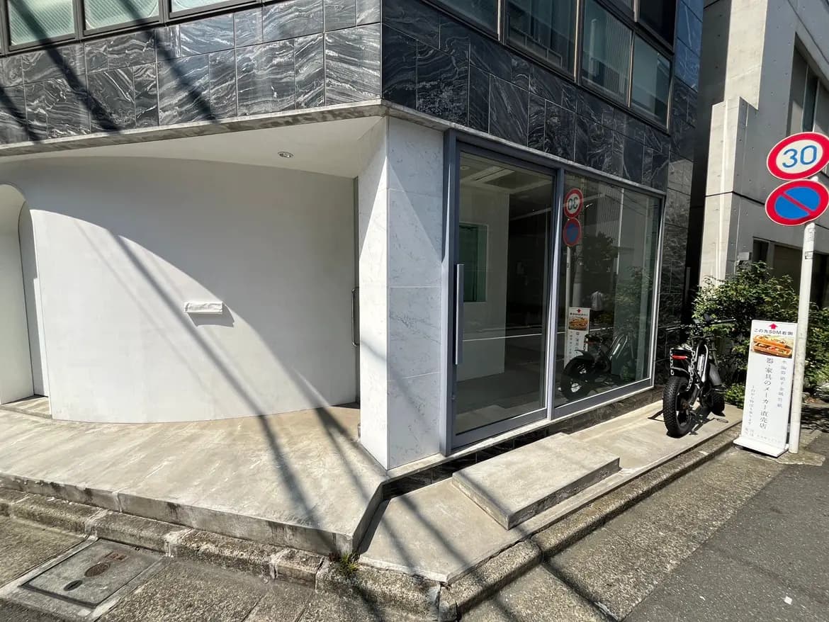 - モダンな建物のエントランス、透明なガラス扉と曲線的なデザインの壁、そばに停まったバイクと交通標識がある外観