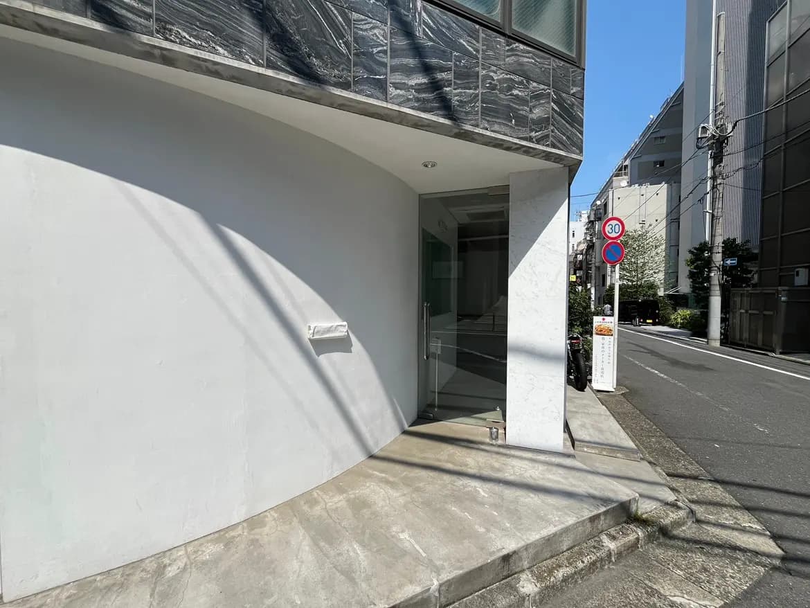- 都市のモダンな建物のエントランス、曲線的な外壁と明るい日差しが差し込む様子、近くには駐車禁止の道路標識が見える