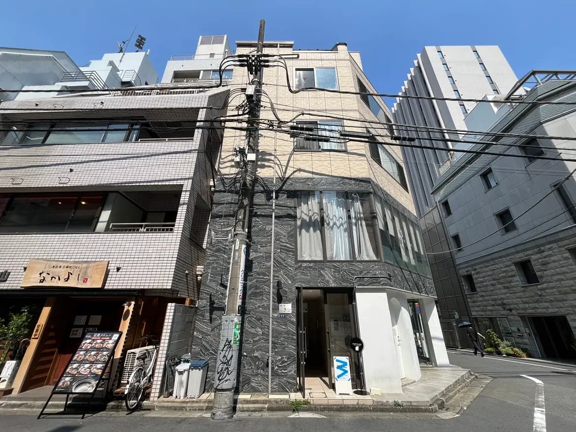 - 都市に建つ現代的なデザインのビル、柱と配線が目立ち、手前には飲食店の看板がある街角の風景