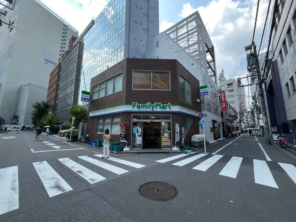 - 都会の交差点に位置するファミリーマートの店舗、周囲には高層ビルと緑が点在し、道を歩く人々が見える光景