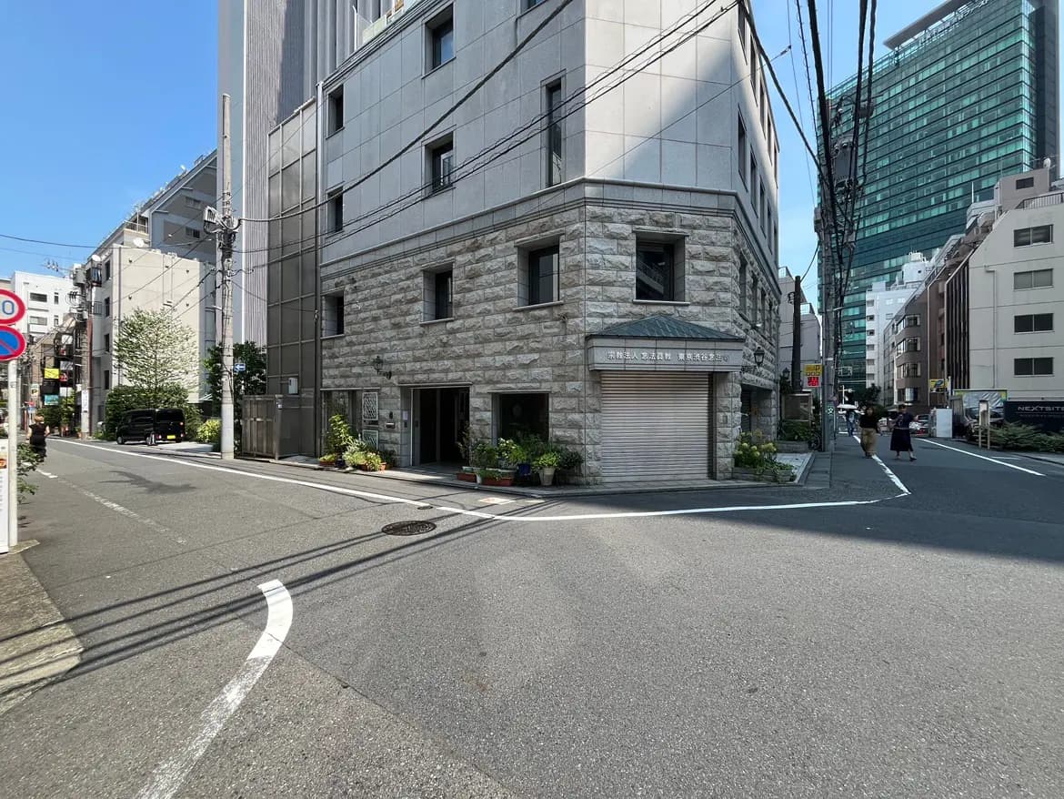 - 都市の角に立つ石造りの建物と近代的な高層ビル、周囲の緑と道路に沿った植栽が特徴的な風景