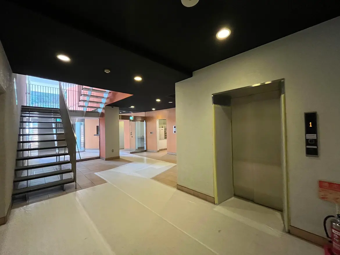- 明るい色合いのロビー、階段とエレベーターがある現代的な建物の内部、床には養生シートが敷かれている