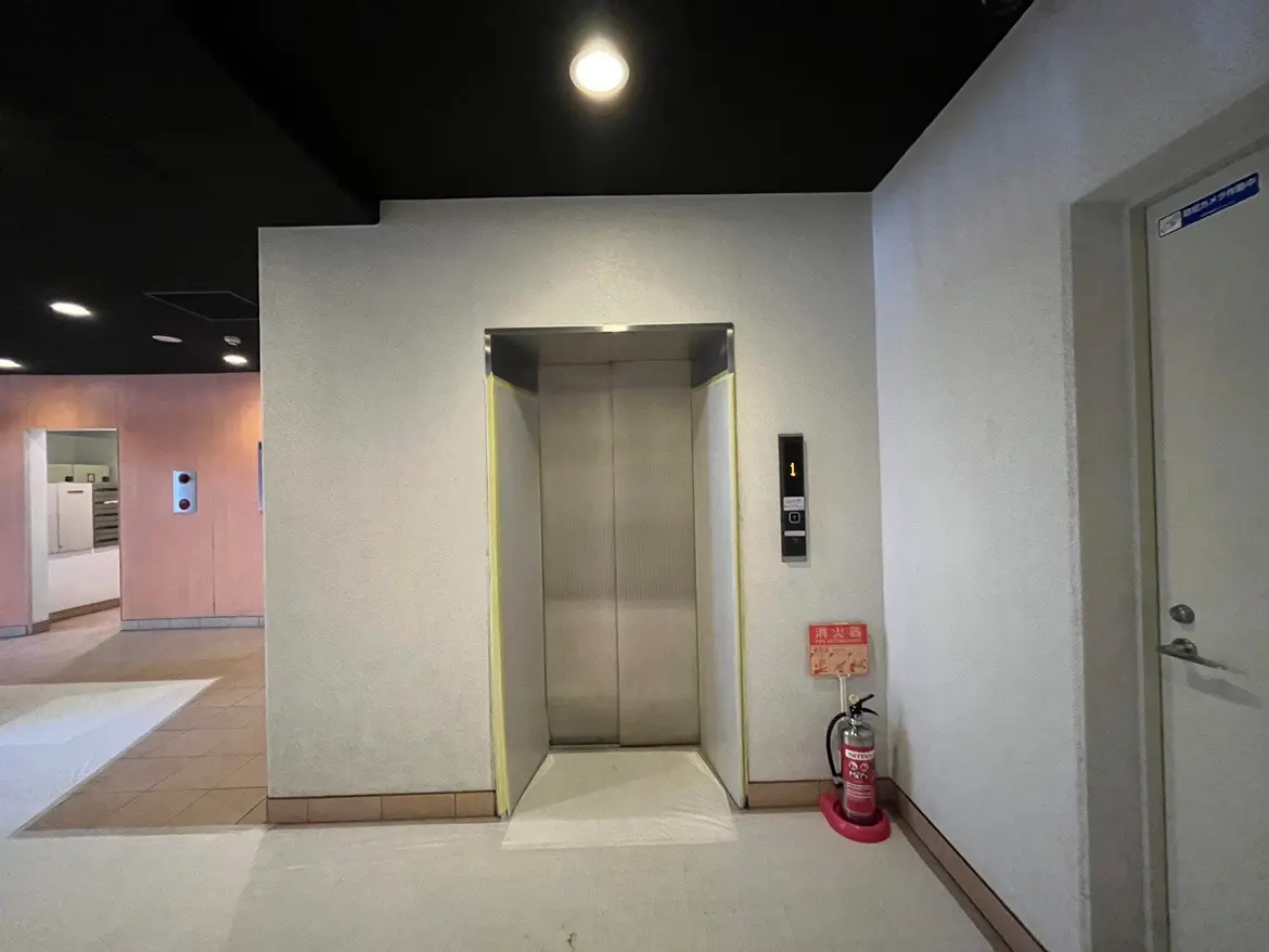 - モダンなビルのエレベーターエントランス、シンプルで清潔感のあるデザイン、火災用消火器が設置されたロビーの様子