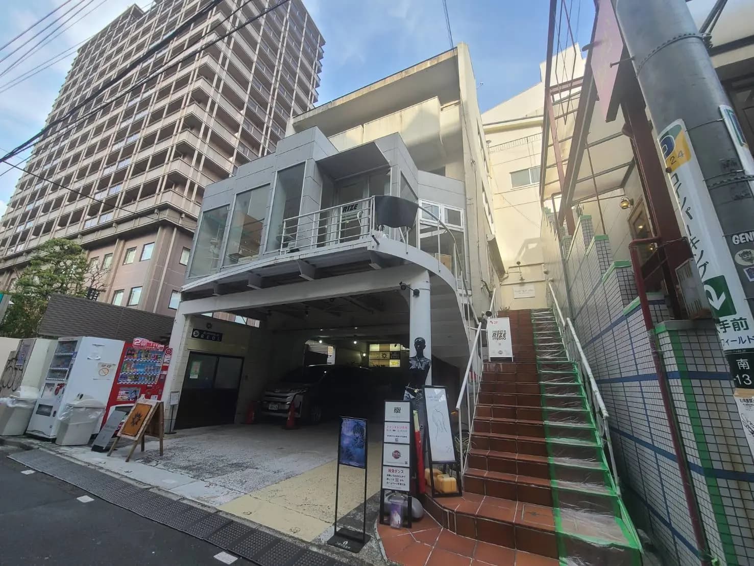 - モダンな外観のビル、ガラスのエントランスと階段、周囲には自動販売機と看板がある街並み