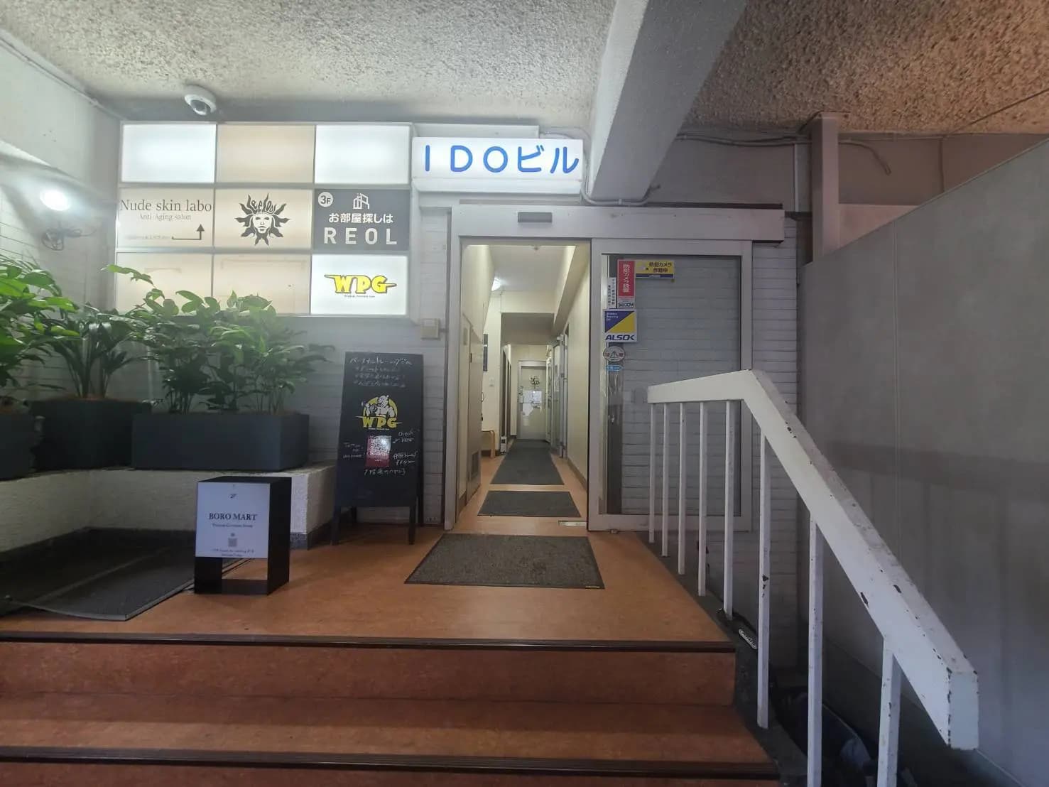 - IDOビルの入り口、看板や観葉植物があり、階段とドアの前にマットが敷かれた様子