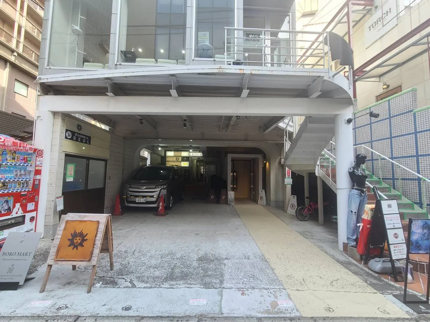 - 商業施設のエントランス、ガラス張りの2階とスペースが広がるアプローチ、右側に展示されている衣料品と自転車駐輪場の案内看板