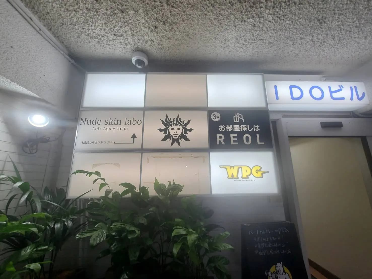 - 植物に囲まれた建物の入り口、看板には「Nude skin labo」や「お部屋探しはREOL」、その他の商業スペースの情報が記載されている様子