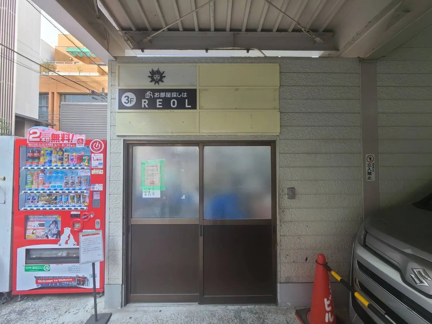 - 自動販売機と出入口があるビルの外観、3階にある「REOL」という看板が見える風景