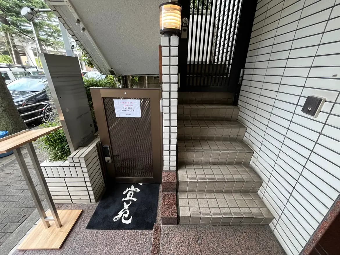 - 階段と入口の様子、モダンなデザインのアプローチ、植栽が施されたエントランスに掲示された営業時間の案内がある