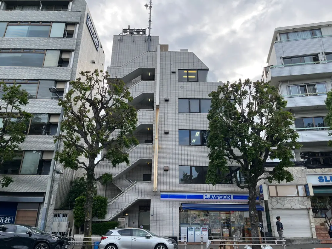 - 都市の街並みに密着した外観のビル、近くにローソンのコンビニエンスストア、緑豊かな樹木が並ぶ風景