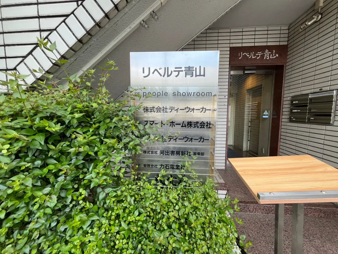 - リベルテ青山の入り口、緑豊かな植栽と企業の看板が並ぶショールームへのアクセスが示されている風景