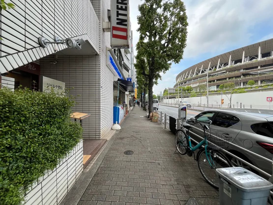 - 都会の歩道沿いに並ぶ店舗とビル、緑豊かな植栽と自転車、背景にはスタジアムが見える風景