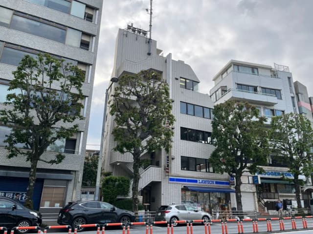 - 駅前のローソンが入る白いタイル張りのビルと周囲の木々、近隣の商業施設が並ぶ街並み