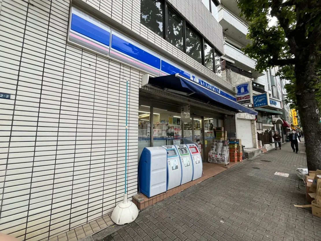 - コンビニエンスストアの外観、青と白の看板、ゴミ箱やATMが並んでいる通りの風景、周囲には歩行者がいる様子