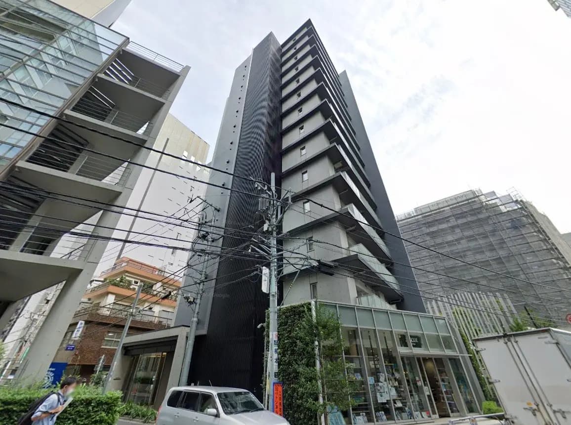 - 周囲のビルに囲まれたスタイリッシュな黒い外観のマンション、近くにはカフェと緑の植栽が見える街並み