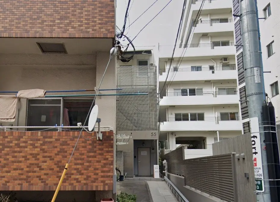 - アパートの外観、隣接する建物と電線が写る都市の風景、洗濯物が干されたバルコニーが特徴