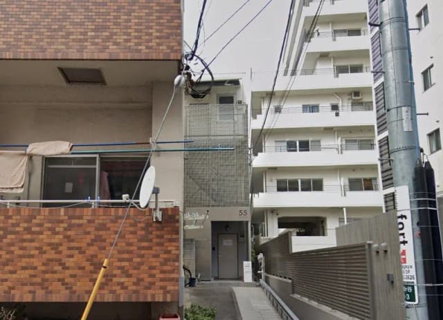 - アパートの外観、隣接する建物と電線が写る都市の風景、洗濯物が干されたバルコニーが特徴