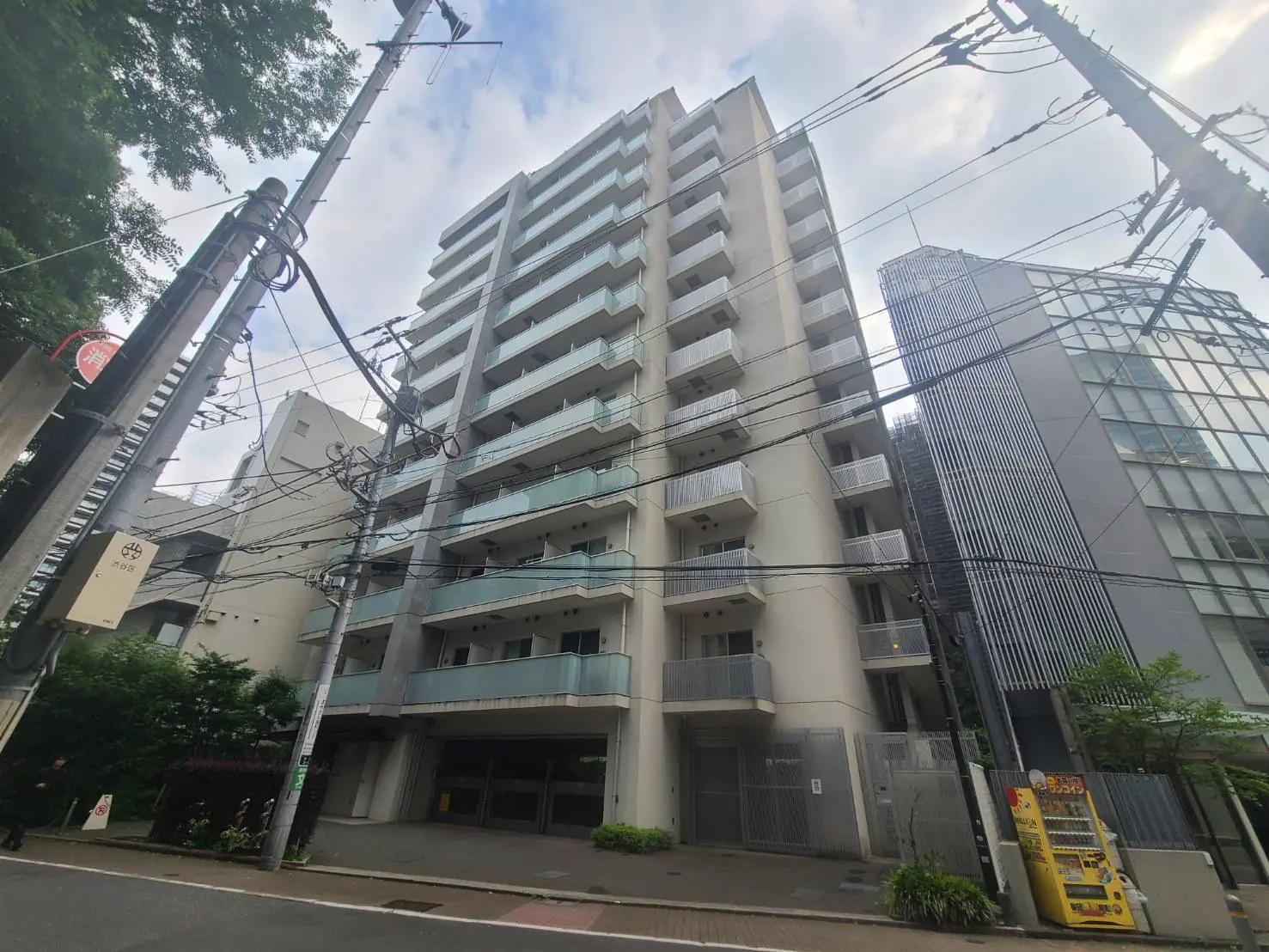 - 都市型マンションの外観、バルコニー付きの階層、周囲には電柱と電線、近代的な建物が立ち並ぶ景観