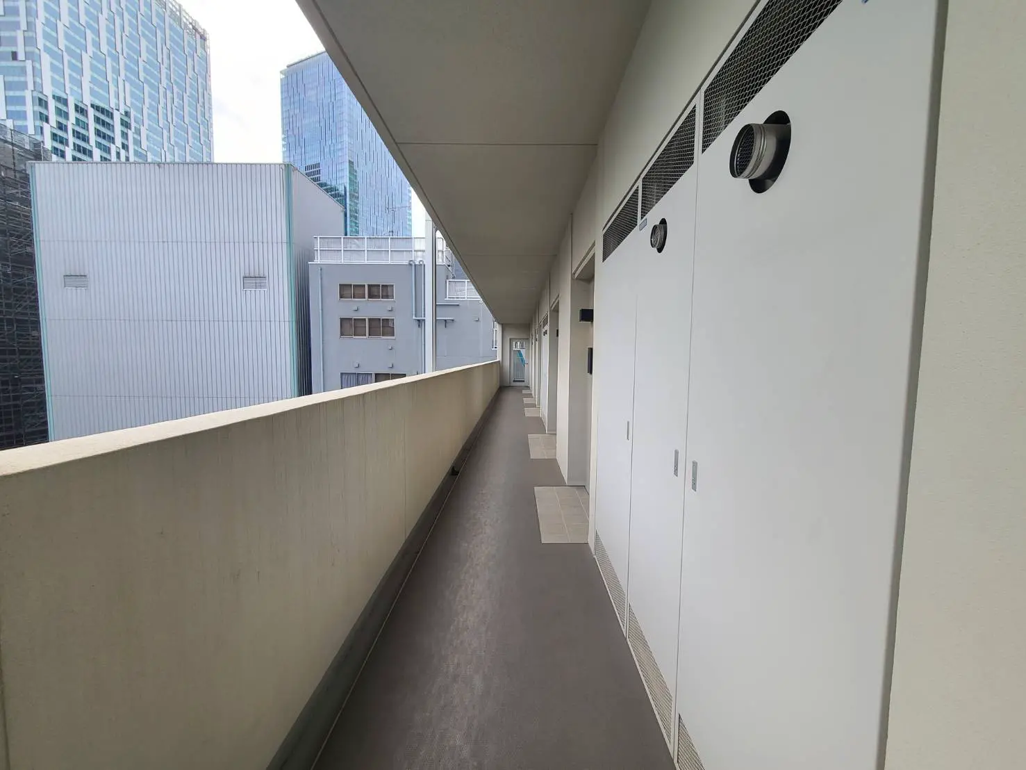 - 都市の高層ビルが立ち並ぶ中の長い廊下、白い壁に扉が並ぶシンプルなデザインの共用スペース