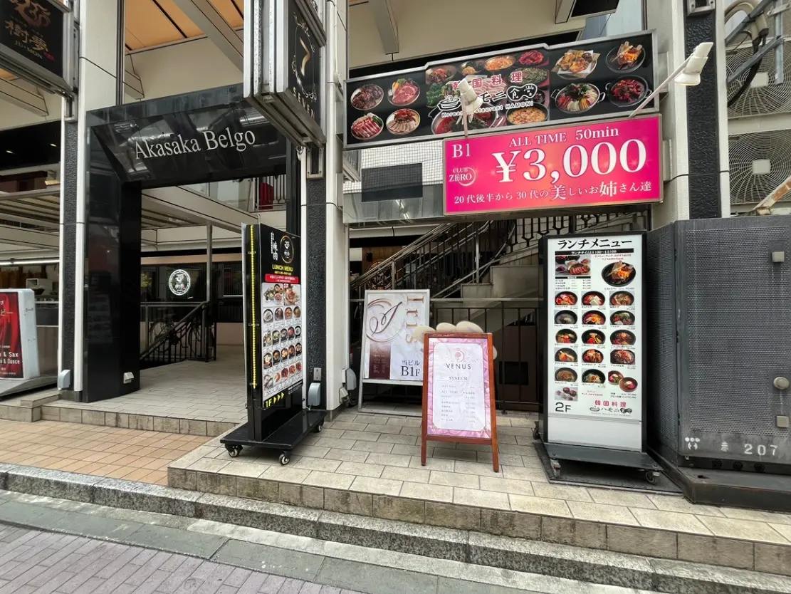 - 赤坂ベルゴの入口、ランチメニューと料金案内が掲示されたボード、周囲の商業施設の外観が映る風景