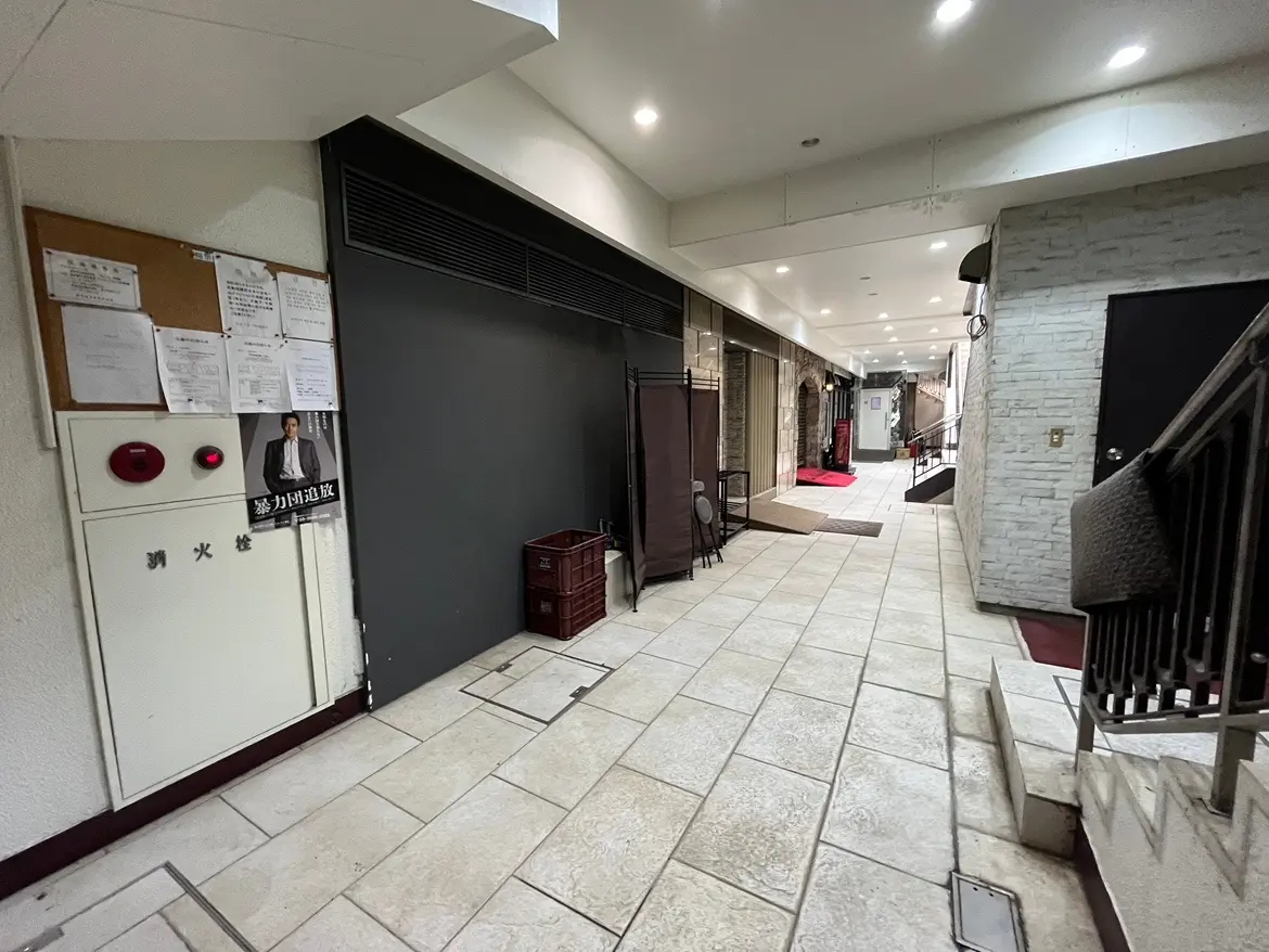 - 厨房や店舗につながる通路、明るい照明とタイル張りの床、掲示板と消火器が設置された現代的なビルの内部