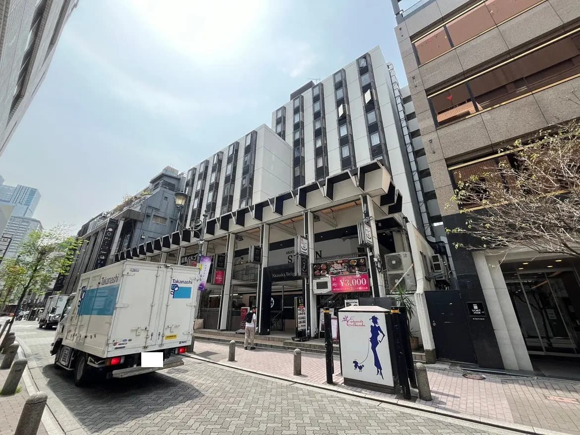 - 都会的な風景、モダンなビル群と配送車が見える通り、店舗看板と歩道に沿った緑の木々