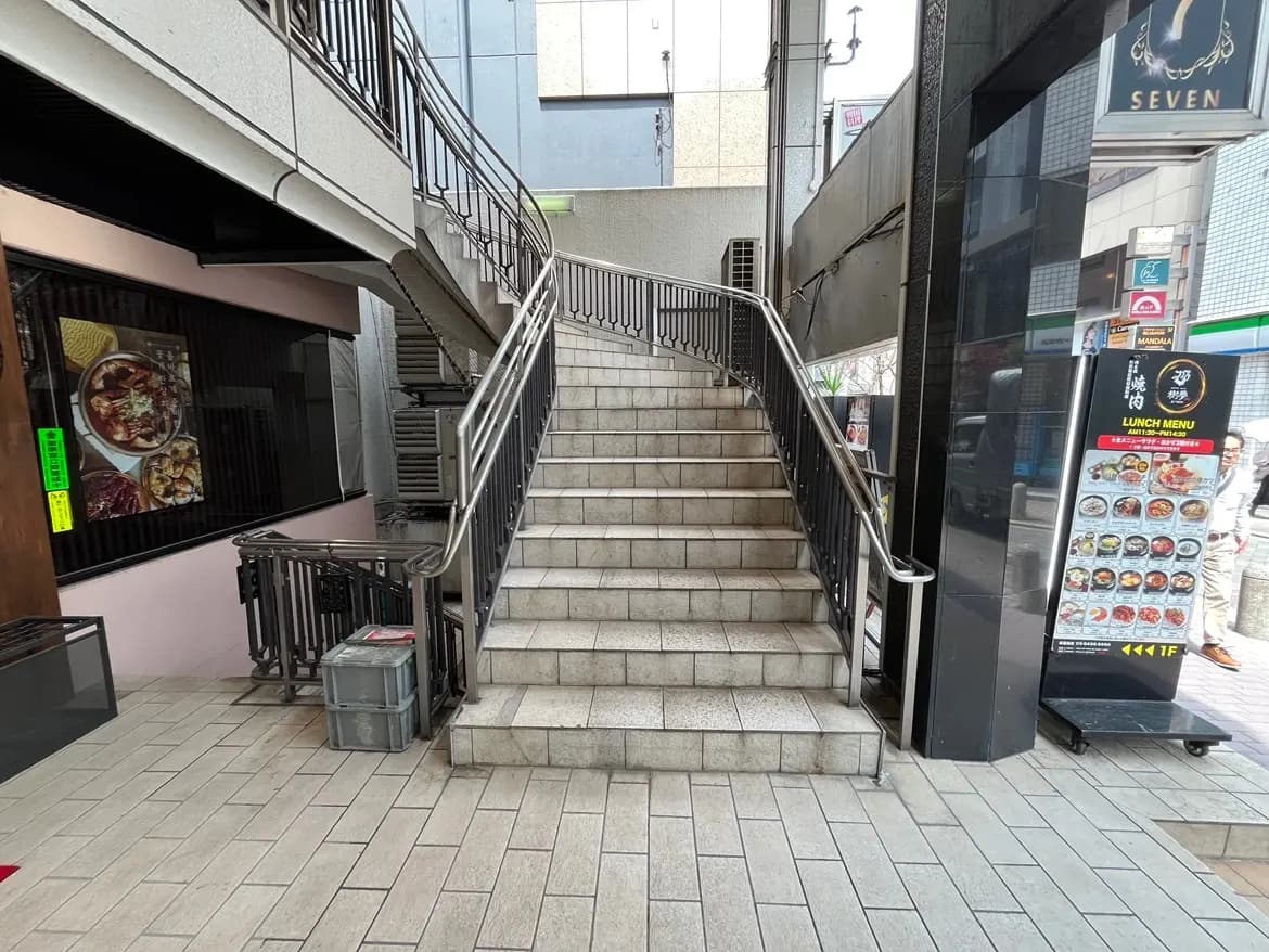 - 屋外の階段とグリル状の手すり、近くの飲食店の看板が見える商業ビルの入口周辺の景観