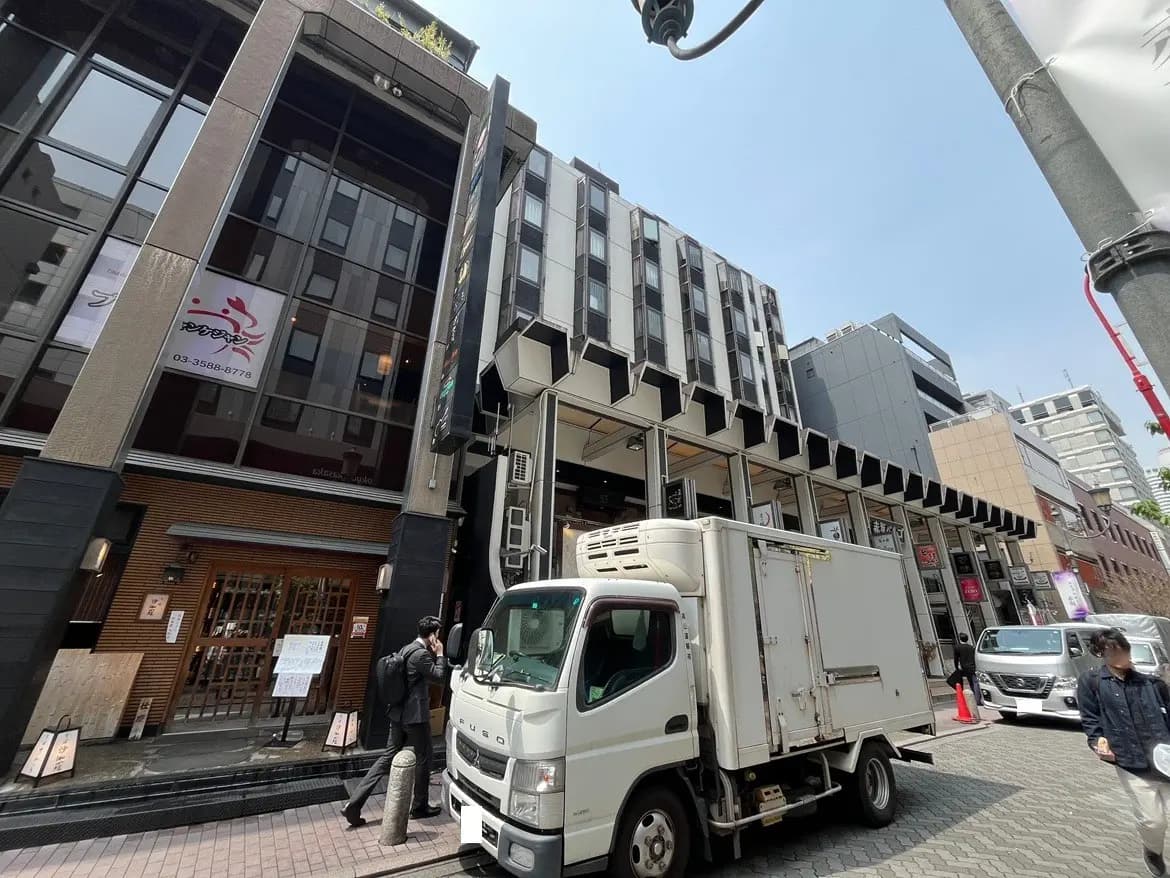 - 商業ビルの外観、ガラスのファサードに広告が掲示され、目の前には白いトラックと通行人がいる繁華街の風景