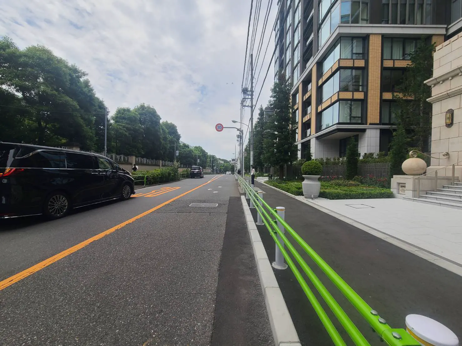 - 大きなマンションが立ち並ぶ景観、車道には黒いミニバンと対向車、緑のガードレールと美しい植栽のある歩道