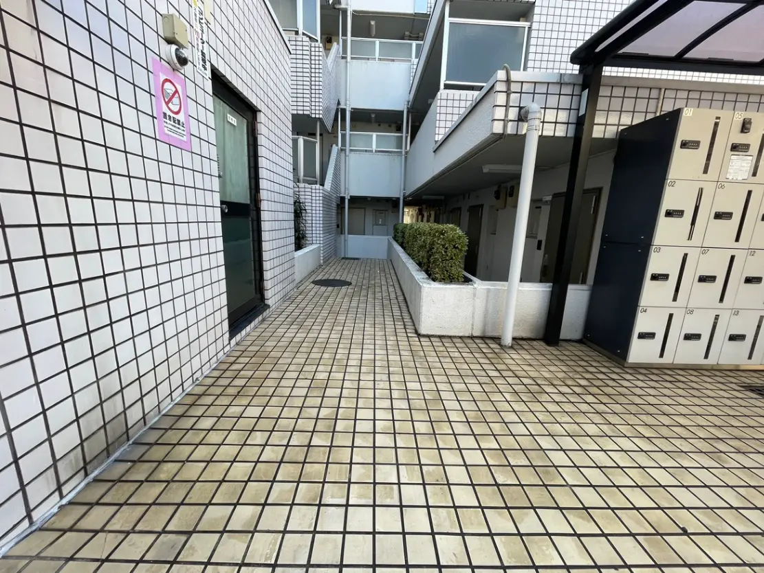 - 清潔感のあるアパートの共用スペース、タイル張りの通路とロッカー、周囲に緑の植栽が施されたエリア