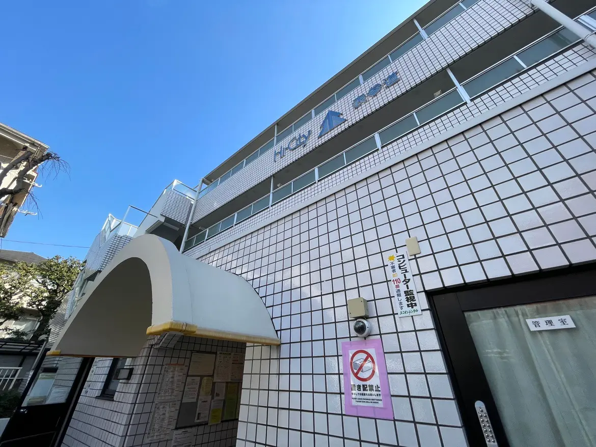 - 白いタイルで覆われたモダンな建物のエントランス、看板が取り付けられた上階、青空を背景にした外観の写真