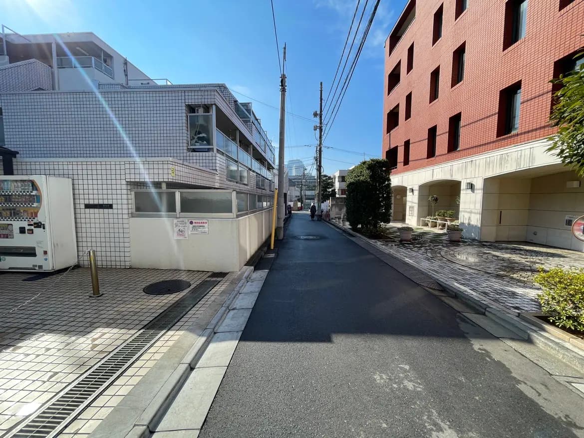 - 明るい青空の下、白いタイルの建物と赤いレンガのマンションが並ぶ静かな通り、周囲には緑の木や花壇が配置された風景