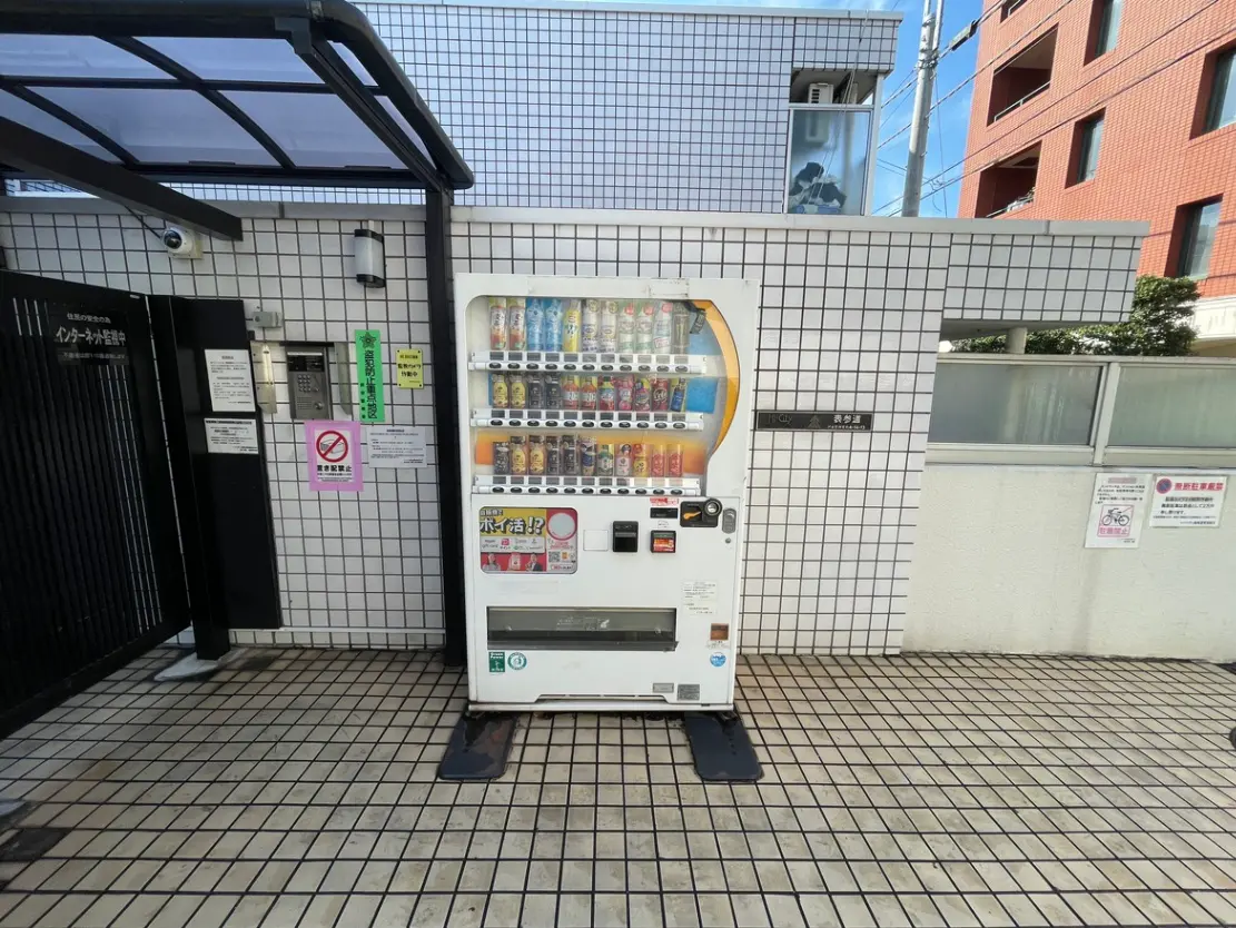 - タイル張りの壁際に設置された飲料自販機、周囲にはインターホンと注意書きが掲示されている風景