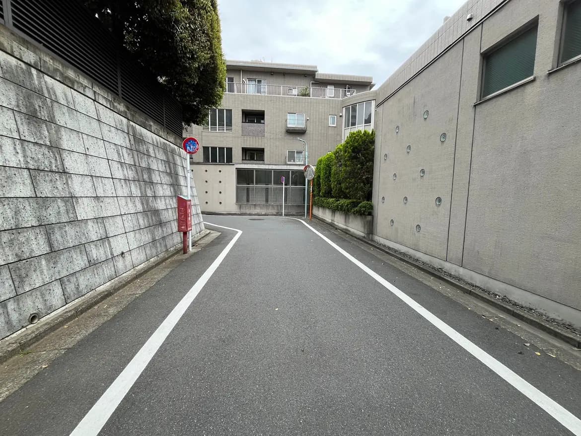 - 都市の細い道路、コンクリートの壁とマンションが立ち並び、交通標識と郵便ポストが見える風景