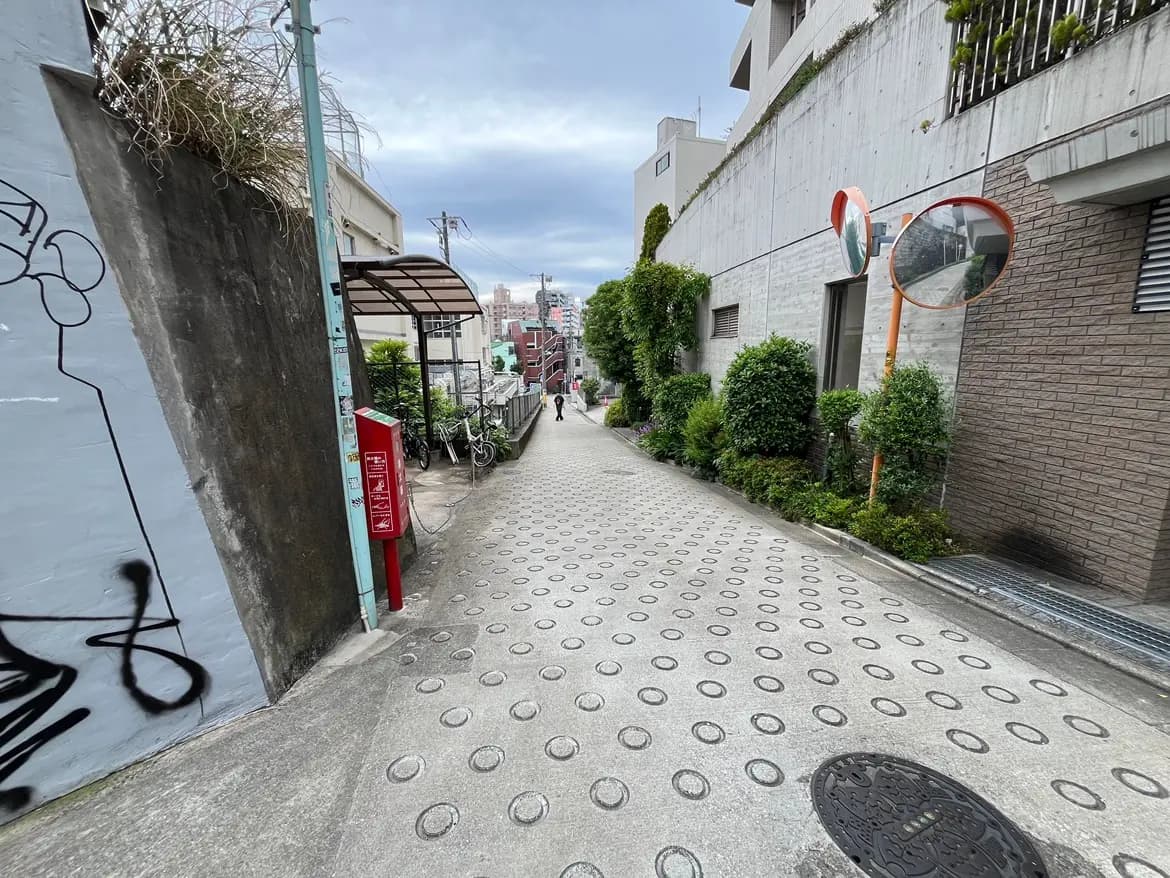 - 都市の狭い道、店舗や住宅が立ち並び、緑の植栽が見える風景、奥にはビルがそびえる情景
