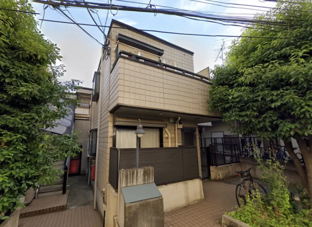- 住宅街にある外観がシンプルな2階建ての家、周囲に緑の植物と自転車が置かれた静かな環境