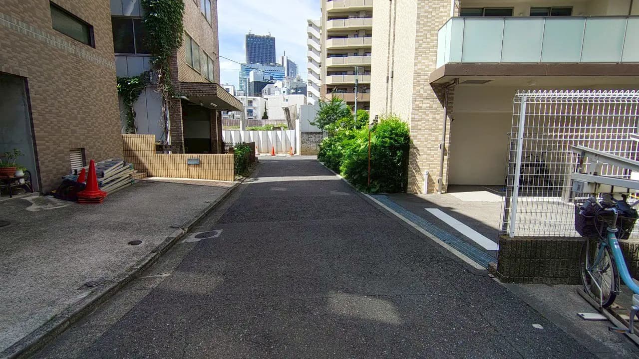 - 都市の住宅街の狭い道路、周囲にはアパートや緑の植栽、遠くにビルや高層建築が見える風景