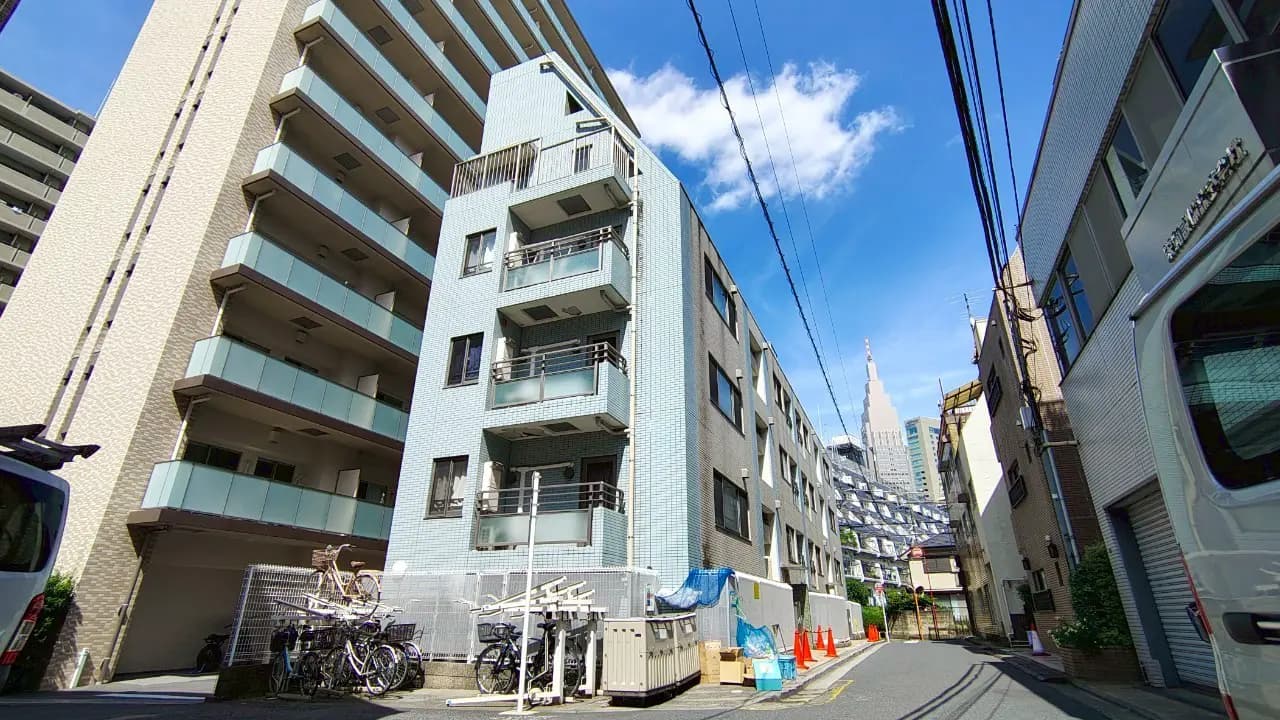 - 都市の住宅街に立つ現代的な低層マンション、青空の下でバルコニーのある外観や周囲のビルと電線が見える風景