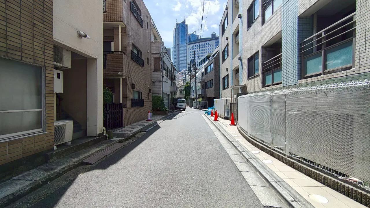- 都市部の狭い通り、両側に建物が並び、青空が見える風景、道路には作業車と赤いコーンが配置されている様子